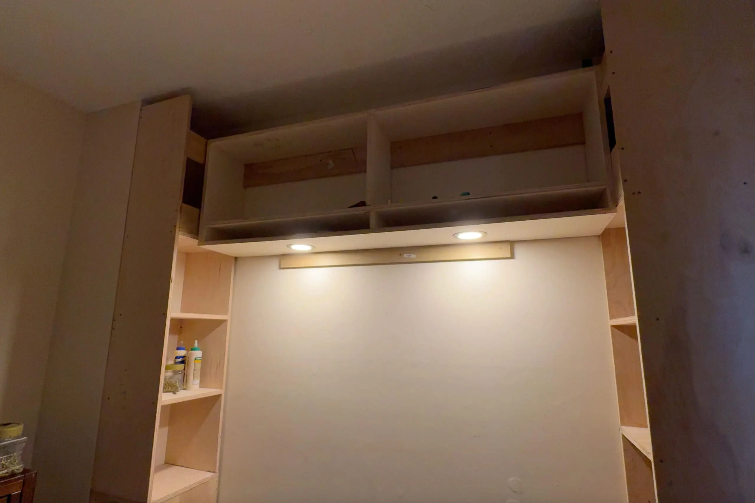 A partially completed DIY built in bedroom unit with recessed lighting installed. The unit consists of two tall plywood storage towers on either side, with open shelving and a central overhead cabinet. Two circular recessed lights are mounted under the top shelf, casting a warm glow on the wall below. Construction tools and materials, including wood glue and screws, are visible on the side shelves, indicating ongoing assembly.